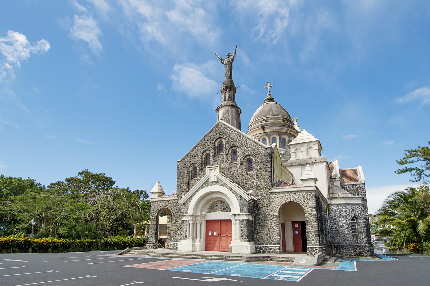 Karibische Kirche aus dunklem Naturstein mit Kuppel und Christusstatue unter blauem Himmel