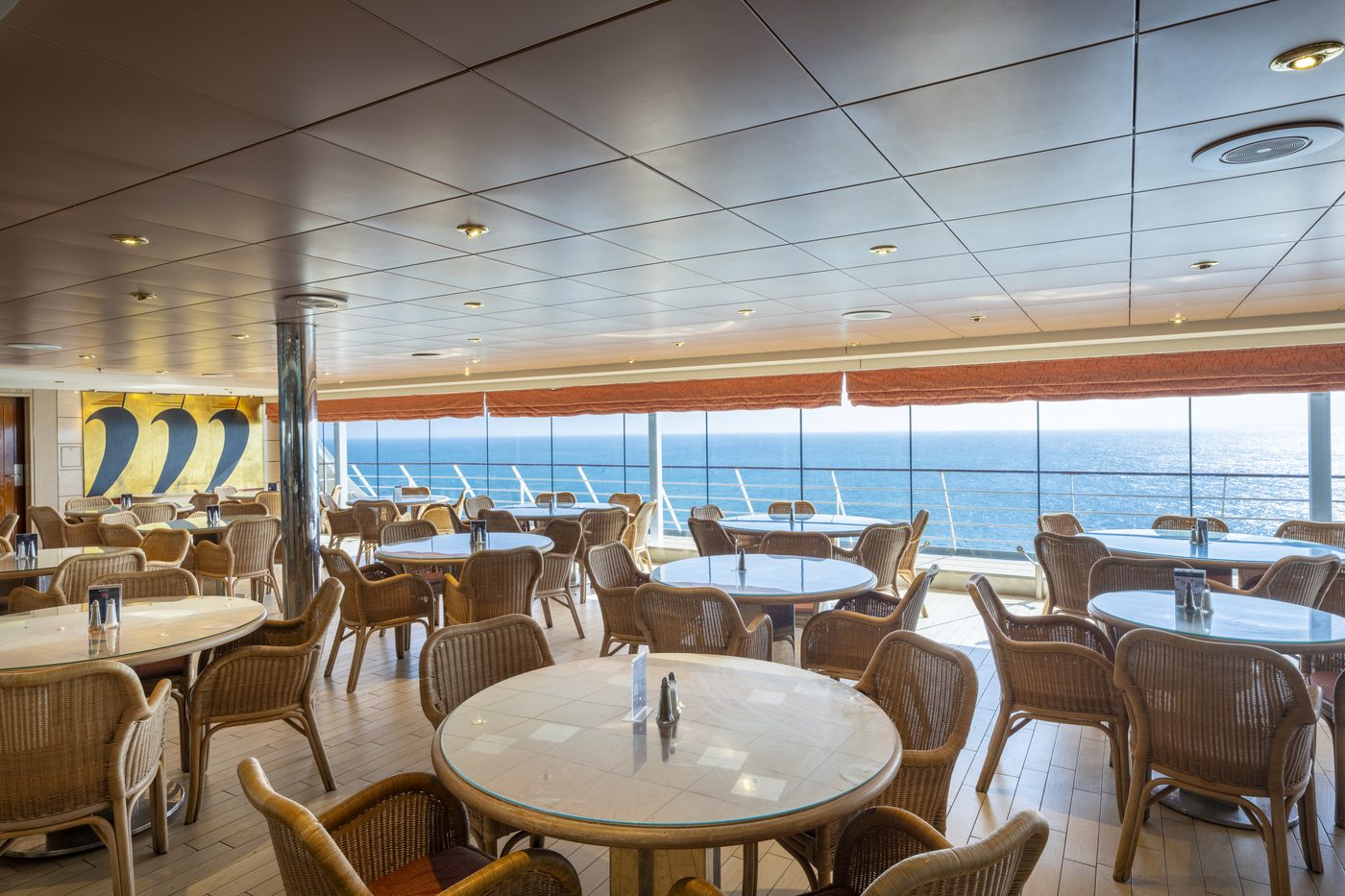 Buffetrestaurant an Bord mit runden Tischen, Korbstühlen und Fensterfront zum Meer