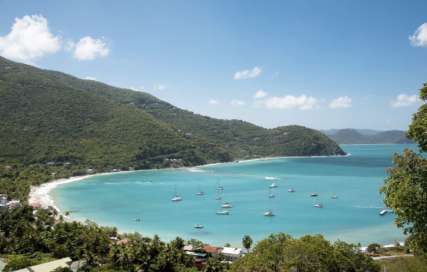 Türkisblaue Bucht mit hellem Sandstrand und ankernden Segelbooten, umrahmt von grünen Hügeln