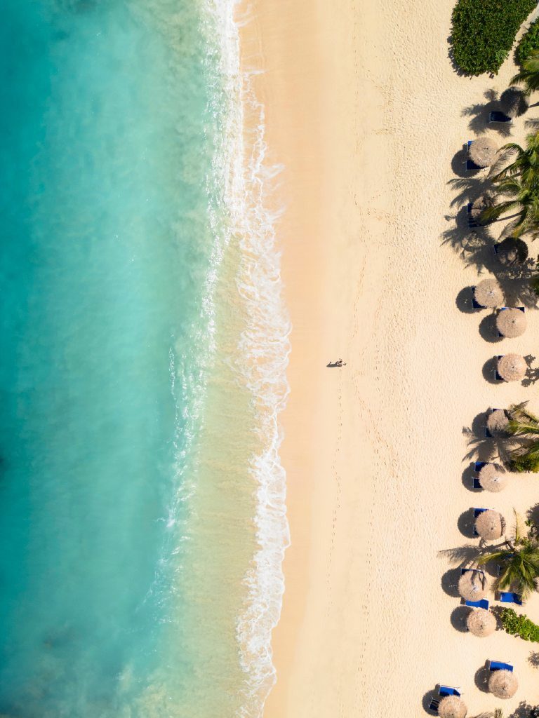 Karibikstrand aus der Vogelperspektive mit türkisblauer Brandung, hellem Sand und Schirmen aus Palmblättern