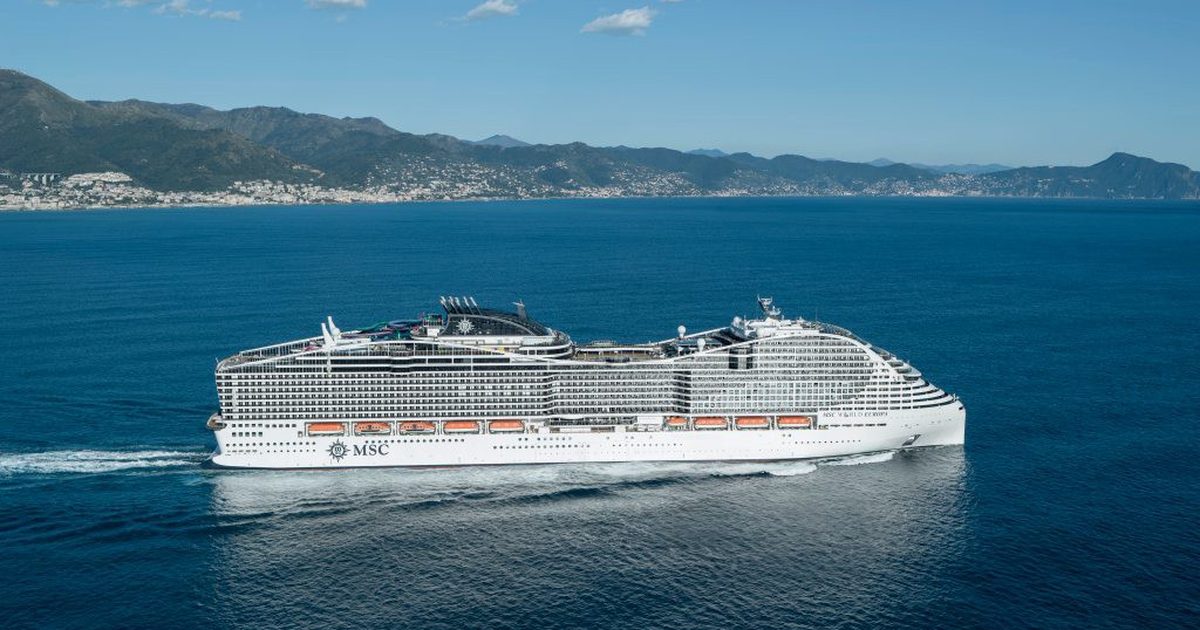 MSC World Europa in voller Seitenansicht vor einer Küstenkulisse mit Bergen, Luftaufnahme im Breitformat